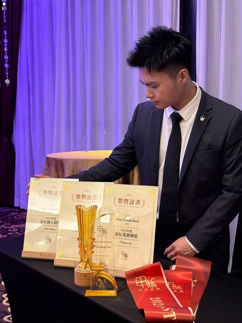 Dexter 穿西装站在桌边，桌上摆着他 2024 年度的荣誉证书和奖杯